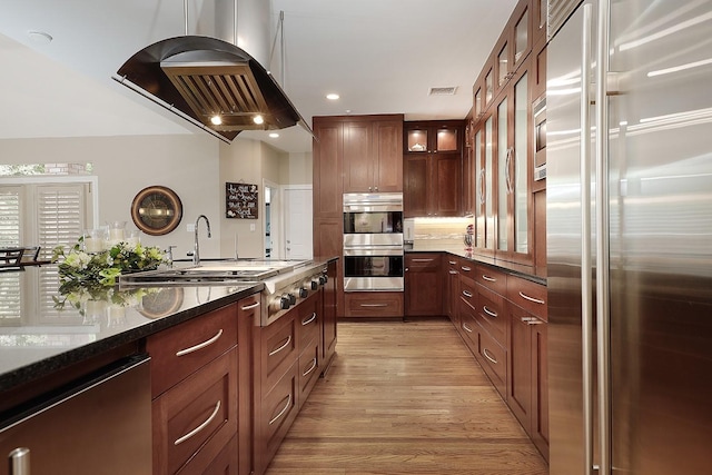 kitchen featuring light wood-type flooring, dark stone counters, stainless steel appliances, island range hood, and hanging light fixtures