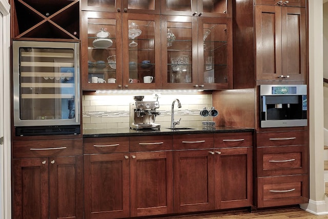 bar featuring decorative backsplash, dark stone counters, stainless steel oven, beverage cooler, and sink