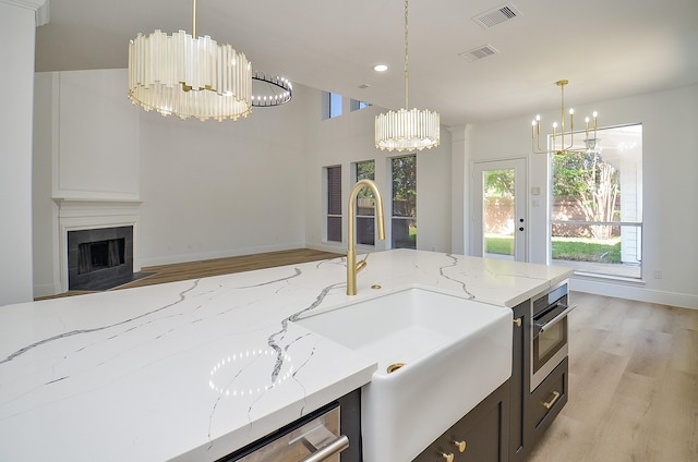 kitchen featuring a tile fireplace, sink, light hardwood / wood-style flooring, decorative light fixtures, and light stone counters