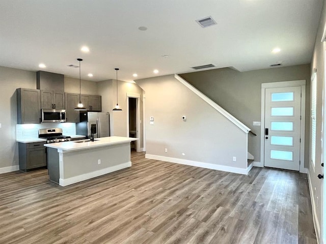 kitchen with gray cabinetry, stainless steel appliances, wood-type flooring, pendant lighting, and a center island with sink