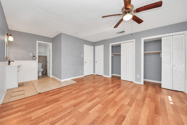 unfurnished bedroom with multiple closets, ceiling fan, ensuite bathroom, a textured ceiling, and light wood-type flooring