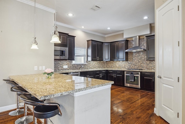 kitchen with kitchen peninsula, appliances with stainless steel finishes, a breakfast bar, wall chimney range hood, and pendant lighting