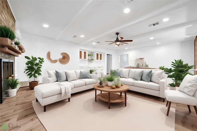 living room with ceiling fan, a stone fireplace, light wood-type flooring, and beam ceiling