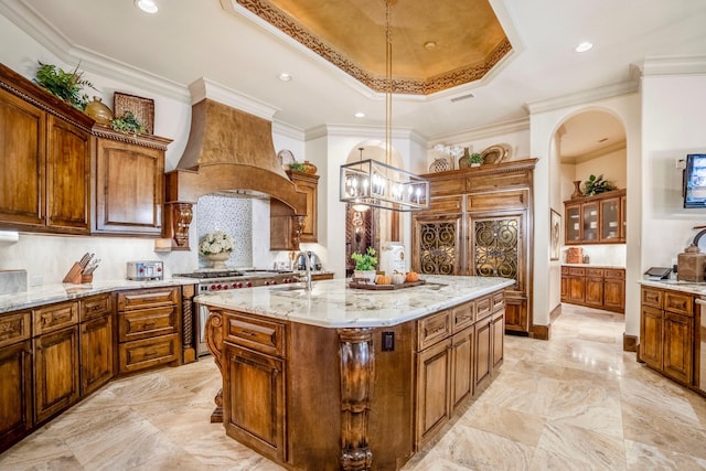 kitchen featuring ornamental molding, custom range hood, a center island with sink, a chandelier, and hanging light fixtures