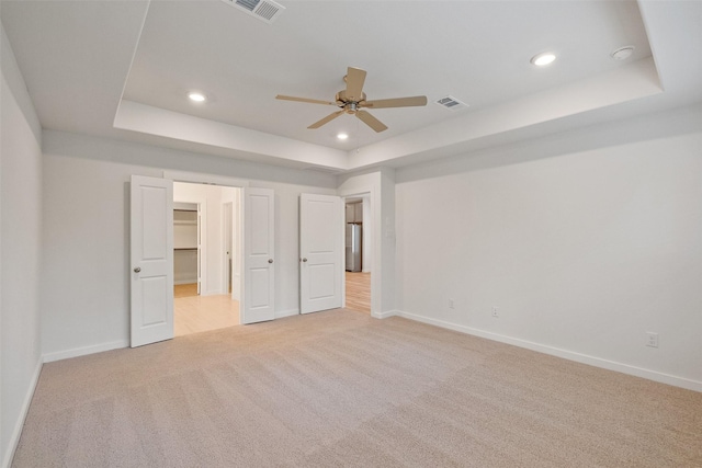 unfurnished bedroom featuring a raised ceiling, a walk in closet, ceiling fan, and light carpet