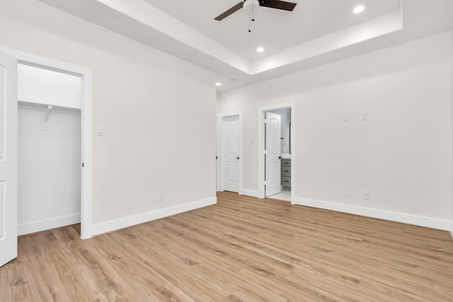 unfurnished bedroom featuring a walk in closet, ensuite bathroom, a raised ceiling, ceiling fan, and light hardwood / wood-style flooring