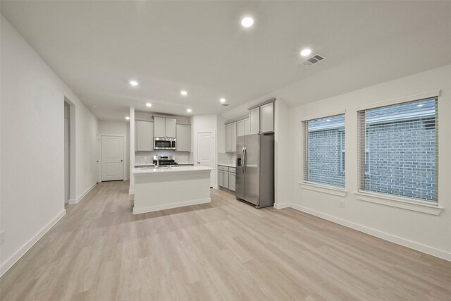 kitchen featuring light hardwood / wood-style flooring, appliances with stainless steel finishes, gray cabinets, an island with sink, and decorative backsplash