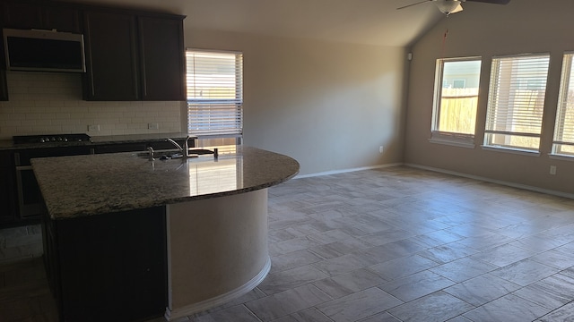 kitchen with backsplash, ceiling fan, sink, a center island with sink, and dark stone countertops