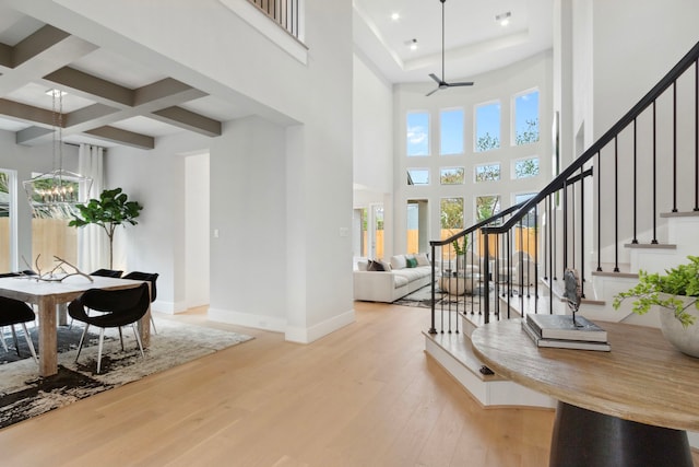 entryway with light wood-type flooring, a towering ceiling, coffered ceiling, ceiling fan with notable chandelier, and beam ceiling