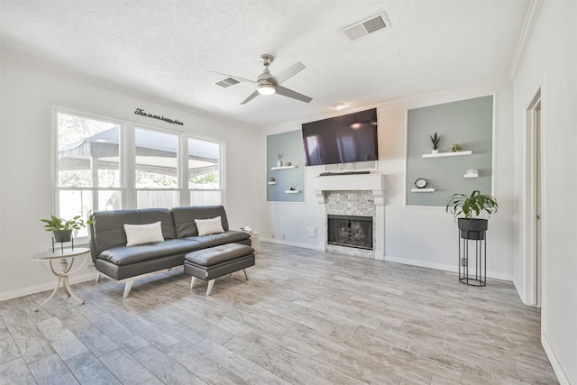 living room featuring ceiling fan, ornamental molding, a textured ceiling, a stone fireplace, and light wood-type flooring