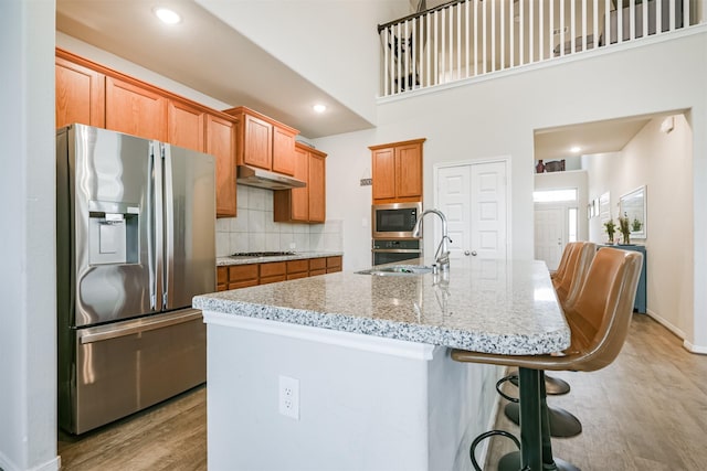 kitchen with appliances with stainless steel finishes, light wood-type flooring, tasteful backsplash, a high ceiling, and an island with sink