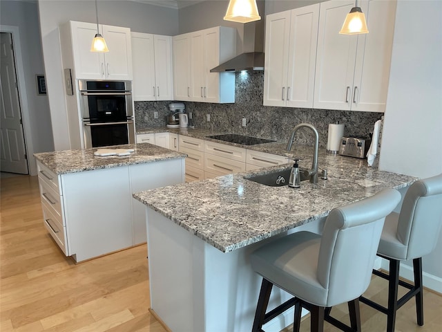 kitchen with kitchen peninsula, wall chimney exhaust hood, stainless steel double oven, white cabinets, and hanging light fixtures