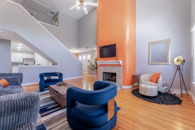 living room with ceiling fan, light wood-type flooring, a towering ceiling, and a tiled fireplace