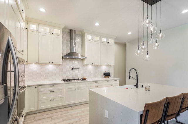 kitchen featuring white cabinets, an island with sink, stainless steel appliances, and wall chimney range hood