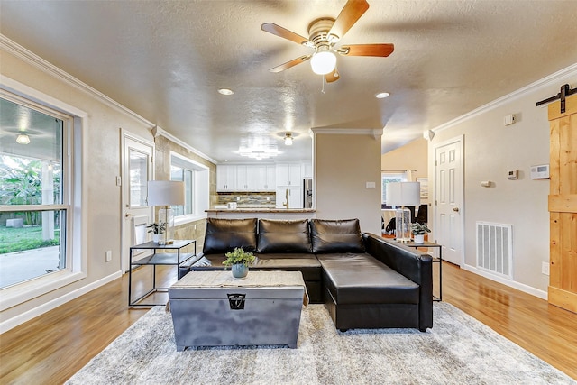 living room featuring ceiling fan, a barn door, light wood-type flooring, ornamental molding, and a textured ceiling