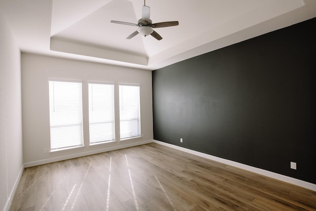 empty room with light wood-type flooring, a tray ceiling, ceiling fan, and baseboards