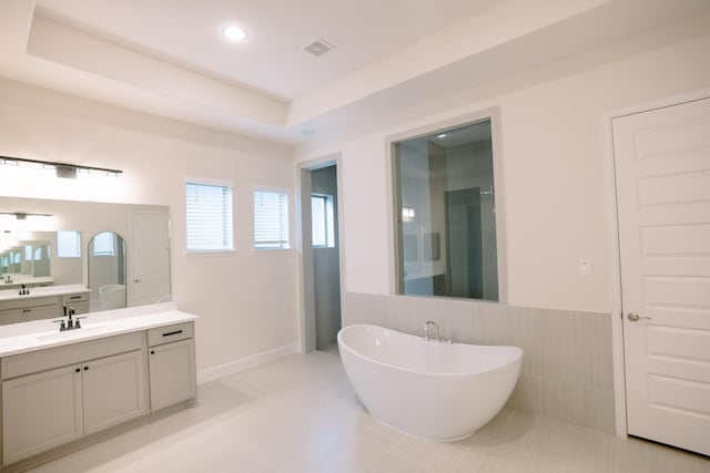 full bath featuring a tray ceiling, vanity, a freestanding tub, tile walls, and recessed lighting