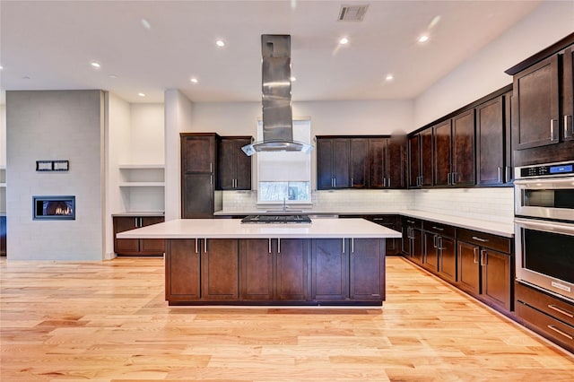 kitchen with appliances with stainless steel finishes, light wood-type flooring, island range hood, a fireplace, and a kitchen island