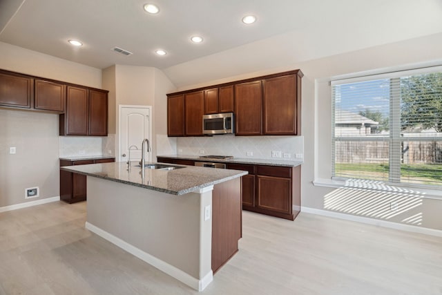 kitchen featuring a kitchen island with sink, sink, vaulted ceiling, light wood-type flooring, and light stone counters