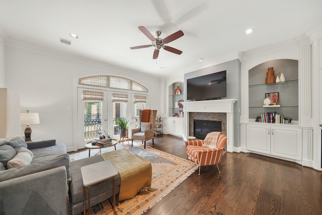 living room featuring built in shelves, ceiling fan, dark hardwood / wood-style flooring, crown molding, and a tiled fireplace