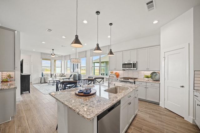 kitchen with appliances with stainless steel finishes, light stone counters, ceiling fan, decorative light fixtures, and a center island with sink