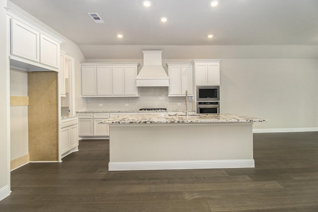 kitchen with custom exhaust hood, built in microwave, a center island with sink, white cabinets, and oven