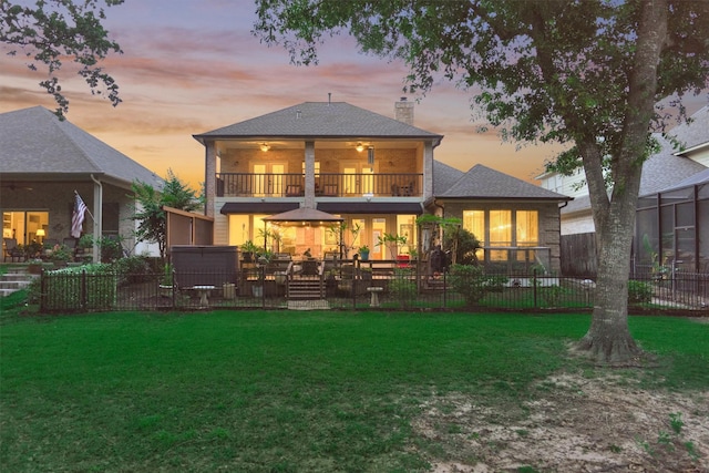 back house at dusk featuring a balcony, ceiling fan, and a lawn