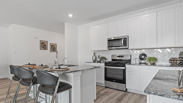 kitchen with stainless steel appliances, dark stone countertops, light hardwood / wood-style floors, a kitchen island with sink, and white cabinets