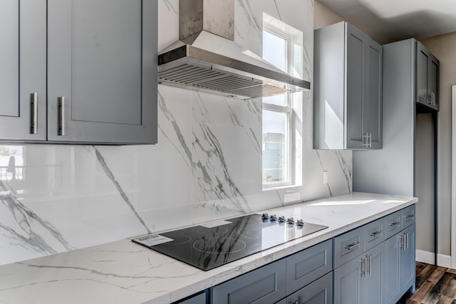 kitchen featuring black electric stovetop, gray cabinetry, wall chimney range hood, and light stone counters