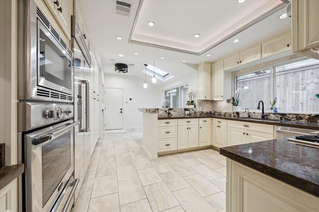 kitchen featuring sink, dark stone countertops, appliances with stainless steel finishes, a raised ceiling, and cream cabinets