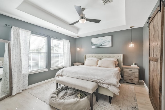 bedroom featuring ceiling fan, a barn door, light hardwood / wood-style flooring, and a tray ceiling