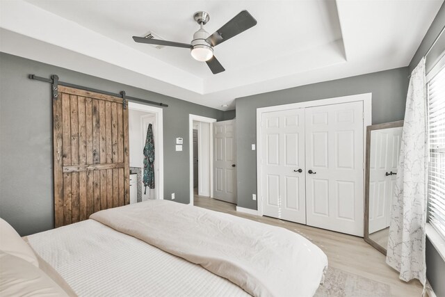 bedroom with a raised ceiling, a barn door, ceiling fan, and light hardwood / wood-style floors