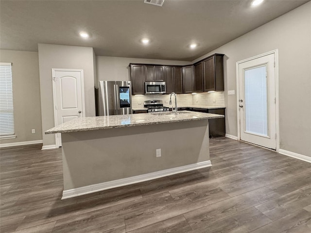 kitchen with light stone countertops, appliances with stainless steel finishes, dark brown cabinets, dark hardwood / wood-style floors, and an island with sink