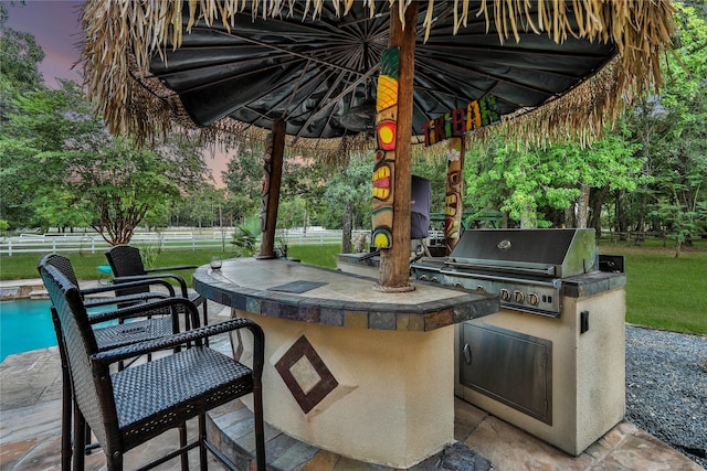 patio terrace at dusk featuring exterior bar, an outdoor kitchen, a fenced in pool, and a lawn