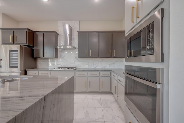 kitchen featuring wall chimney exhaust hood, gray cabinets, tasteful backsplash, light stone counters, and stainless steel appliances