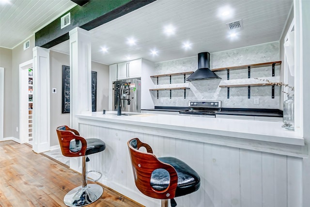 kitchen featuring a breakfast bar, white cabinets, wall chimney exhaust hood, light wood-type flooring, and appliances with stainless steel finishes