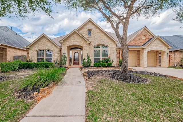 french country home featuring an attached garage, brick siding, a shingled roof, concrete driveway, and a front yard