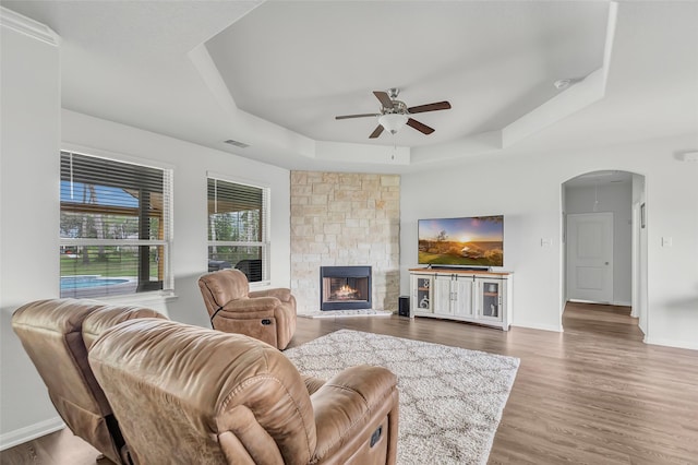 living room with a tray ceiling, a stone fireplace, ceiling fan, and hardwood / wood-style floors