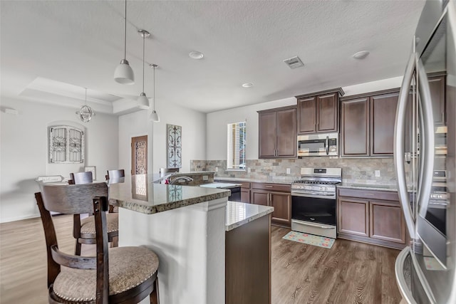 kitchen with light stone countertops, stainless steel appliances, decorative light fixtures, a tray ceiling, and a kitchen bar