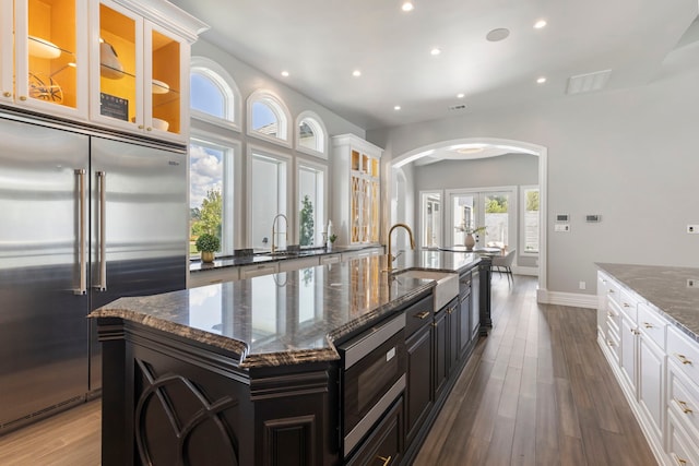 kitchen featuring sink, built in appliances, dark stone countertops, a center island with sink, and white cabinets