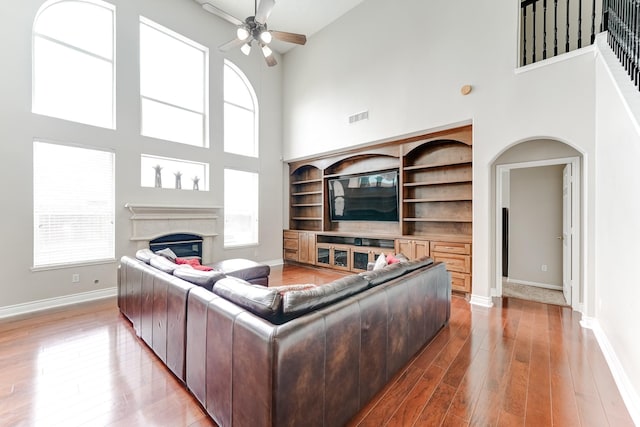 living room featuring built in shelves, ceiling fan, wood-type flooring, and a towering ceiling