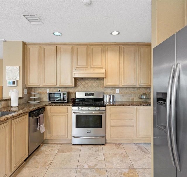 kitchen with appliances with stainless steel finishes, light brown cabinets, backsplash, and dark stone countertops