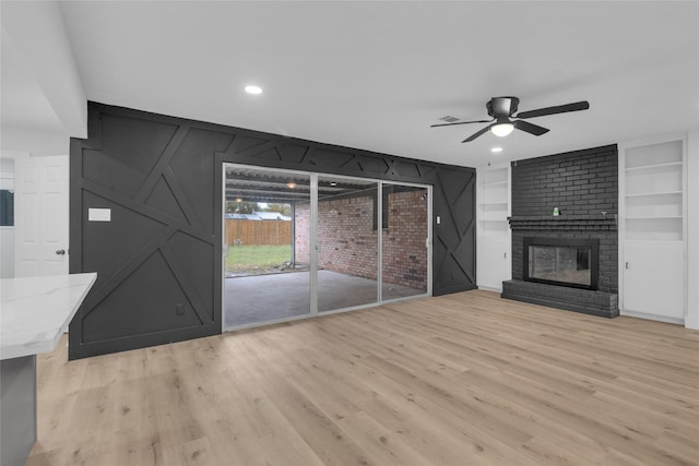 unfurnished living room with ceiling fan, built in features, light wood-type flooring, and a brick fireplace