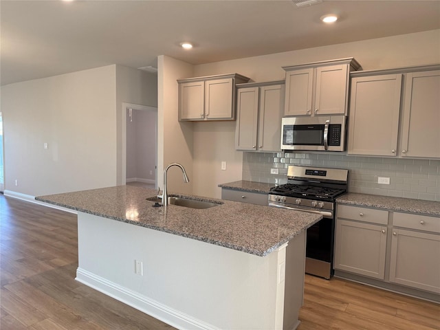 kitchen with light wood-type flooring, stainless steel appliances, sink, stone countertops, and a center island with sink