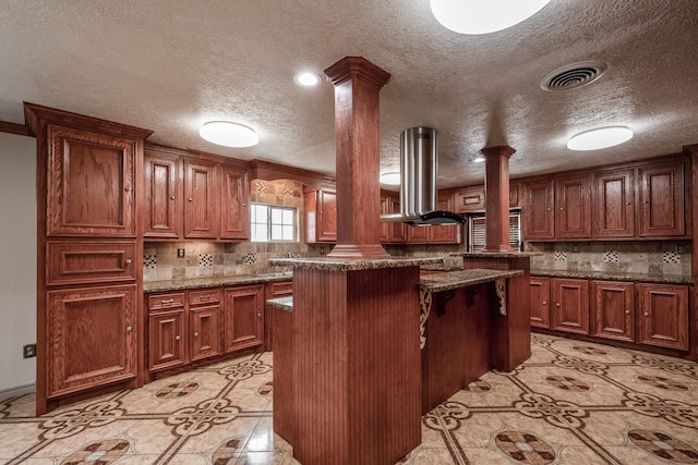 kitchen featuring a kitchen bar, decorative columns, dark stone counters, island range hood, and a center island