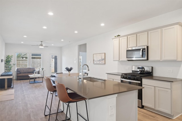 kitchen with light wood finished floors, a kitchen island with sink, a sink, stainless steel appliances, and dark countertops