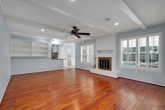 unfurnished living room with a brick fireplace, a textured ceiling, ceiling fan, hardwood / wood-style flooring, and beamed ceiling