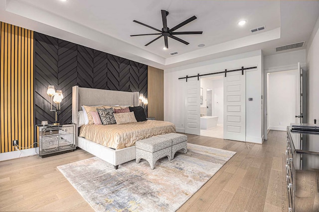 bedroom featuring ensuite bath, a raised ceiling, light wood-type flooring, ceiling fan, and a barn door