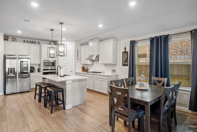 kitchen featuring appliances with stainless steel finishes, a kitchen island with sink, light hardwood / wood-style floors, white cabinetry, and hanging light fixtures