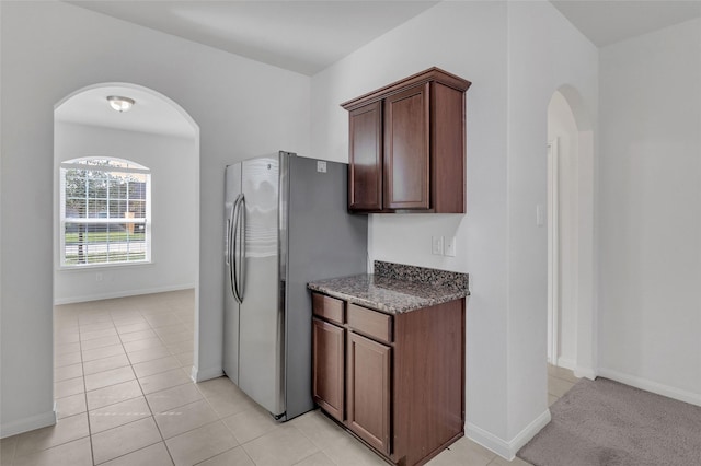 kitchen with light tile patterned flooring, stainless steel fridge, dark brown cabinetry, and dark stone countertops